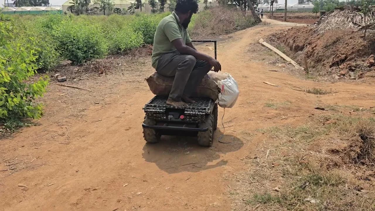 A man sitting on the Versor robot as it carries him and a sack along a dirt track on a farm